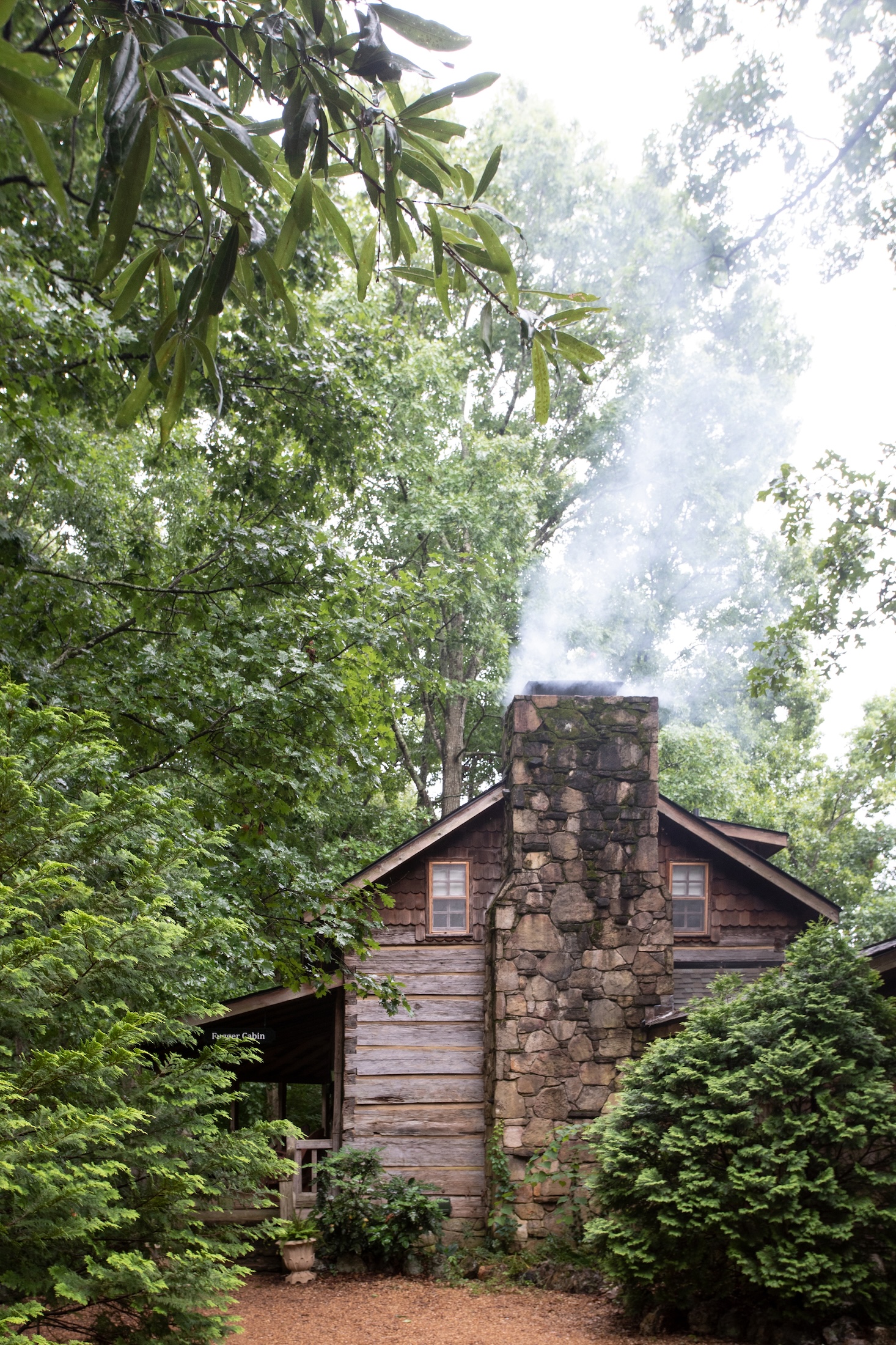 The wooden Fugger Cabin with smoke coming out of the stone chimney sits amongst large, lush, green trees at Barnsley Resort.