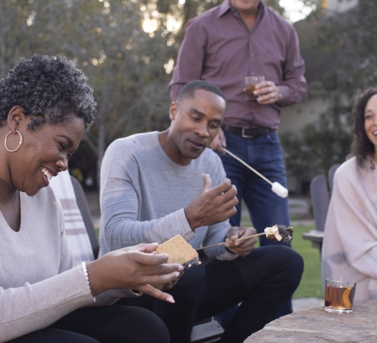 Four people sitting around a stone fire pit making s'mores, having drinks and smiling at Barnsley Resort.