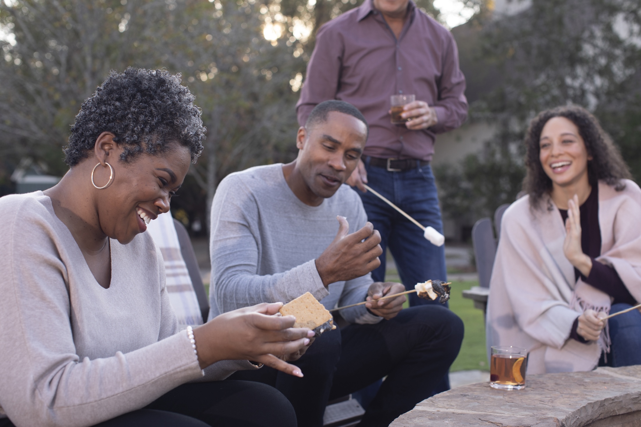 Four people sitting around a stone fire pit making s'mores, having drinks and smiling at Barnsley Resort.