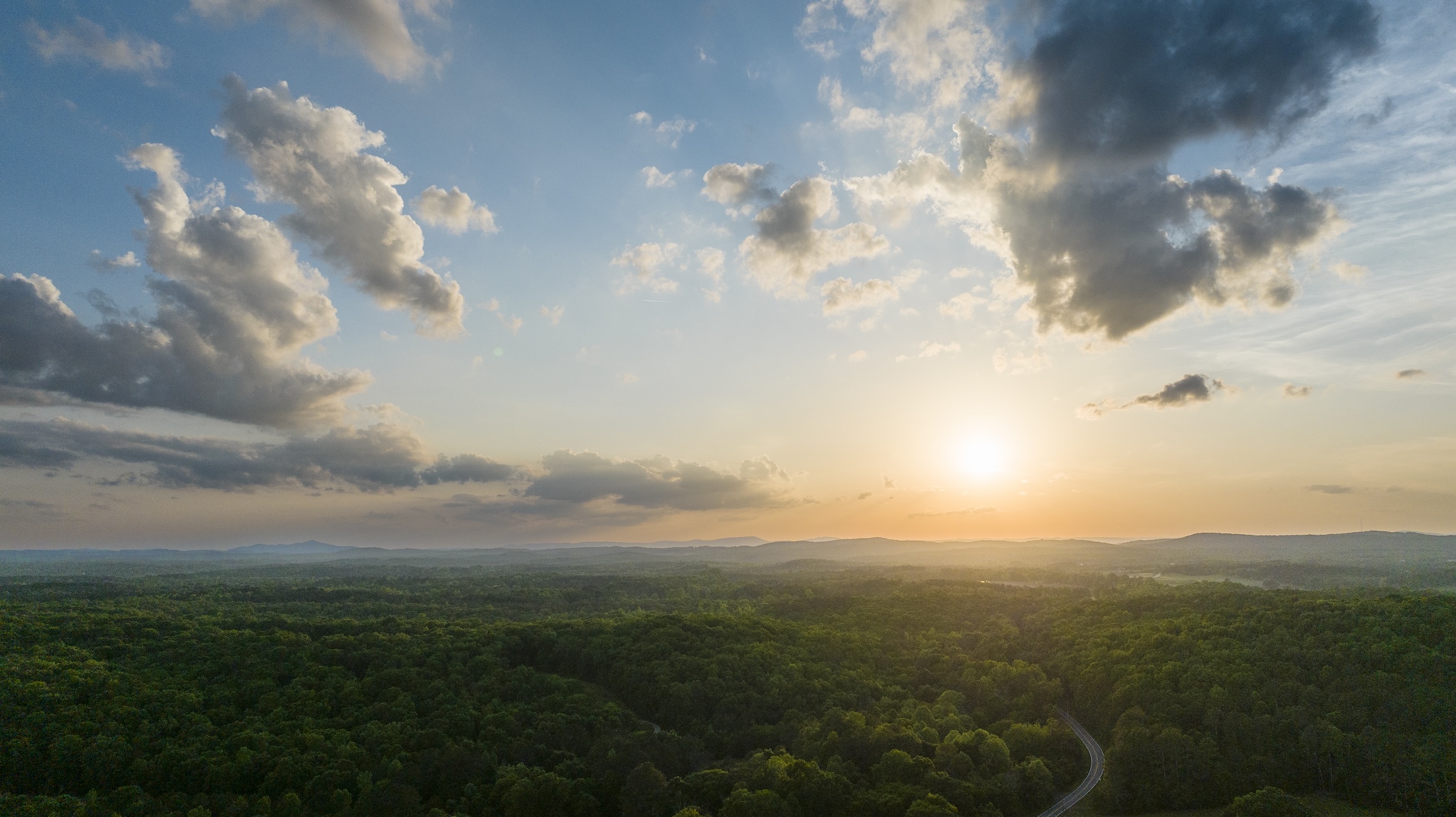 An aerial view of lush forest and a sunset in the distance.