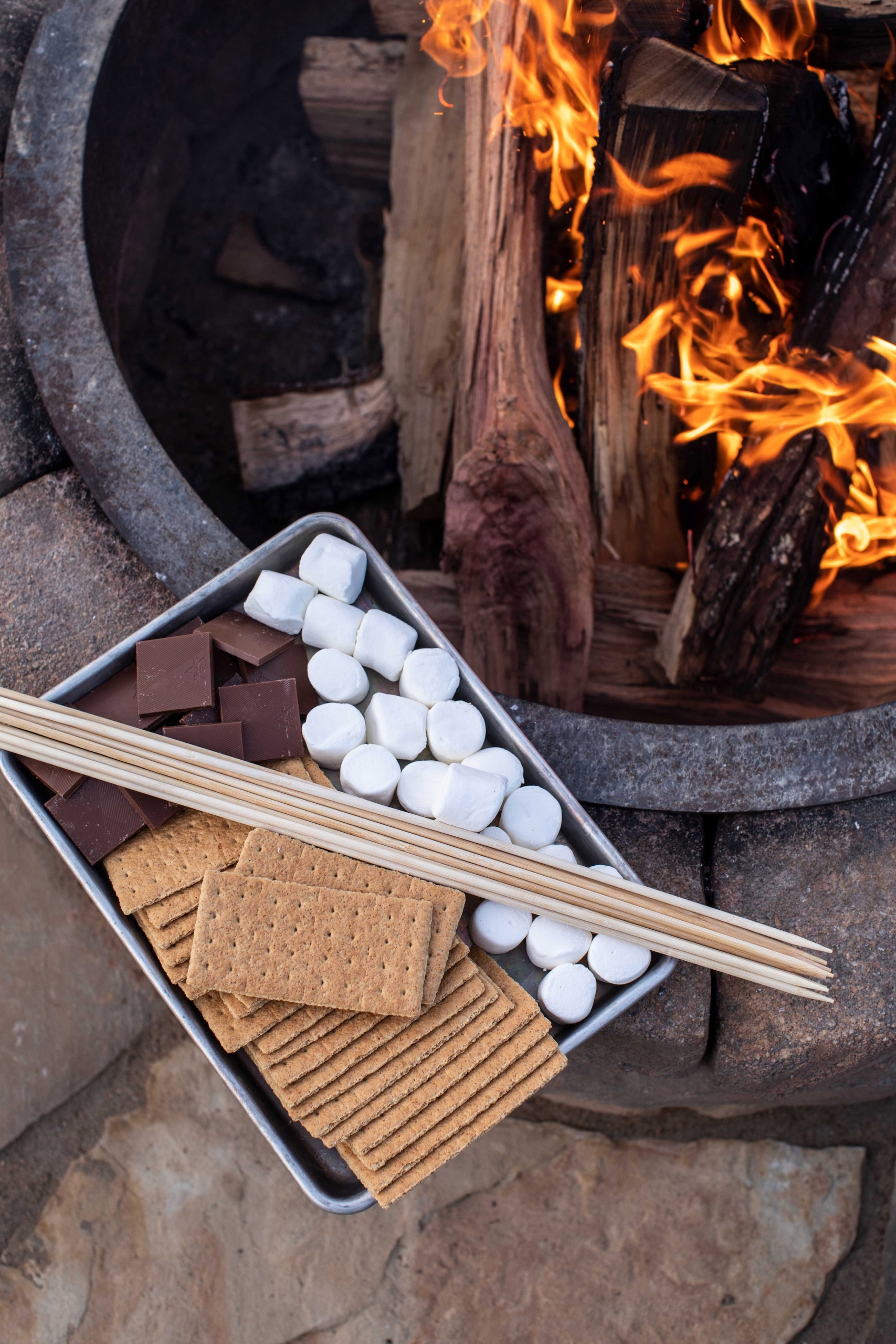 Marshmallows, graham crackers, chocolate and skewers next to a stone fire pit.