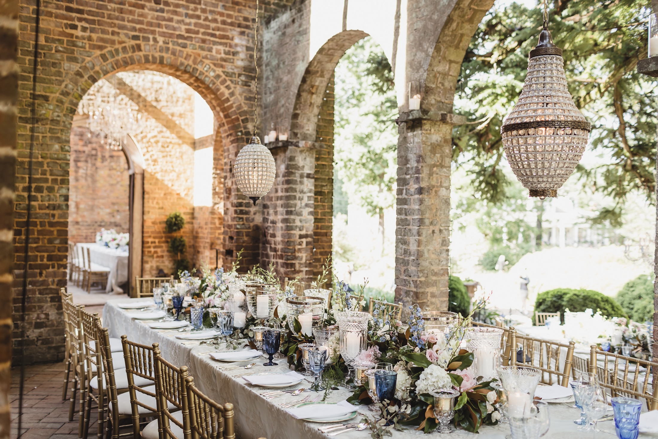 An banquet table set up for a wedding in an open stone building with an ornate chandelier hanging from the branches.