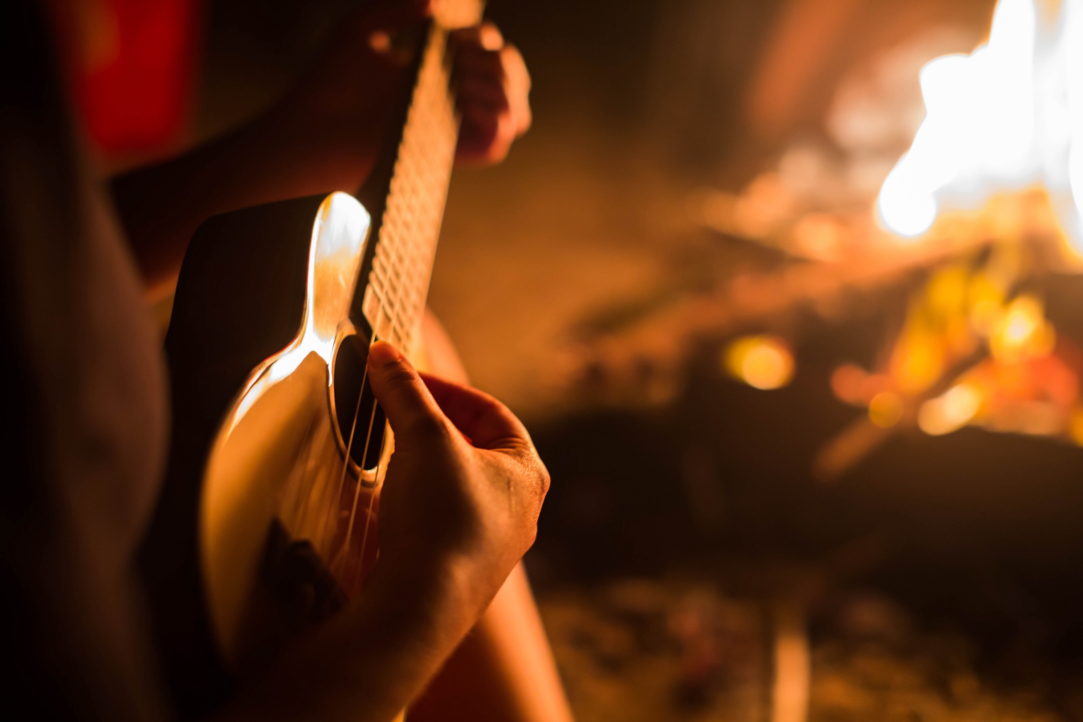 A female musician playing guitar outside, sitting next to a fire.