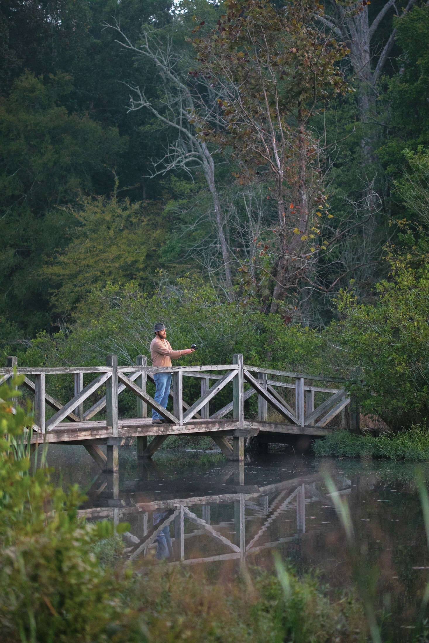 A man fishing from a small walking bridge over a still body of water on a fall morning.