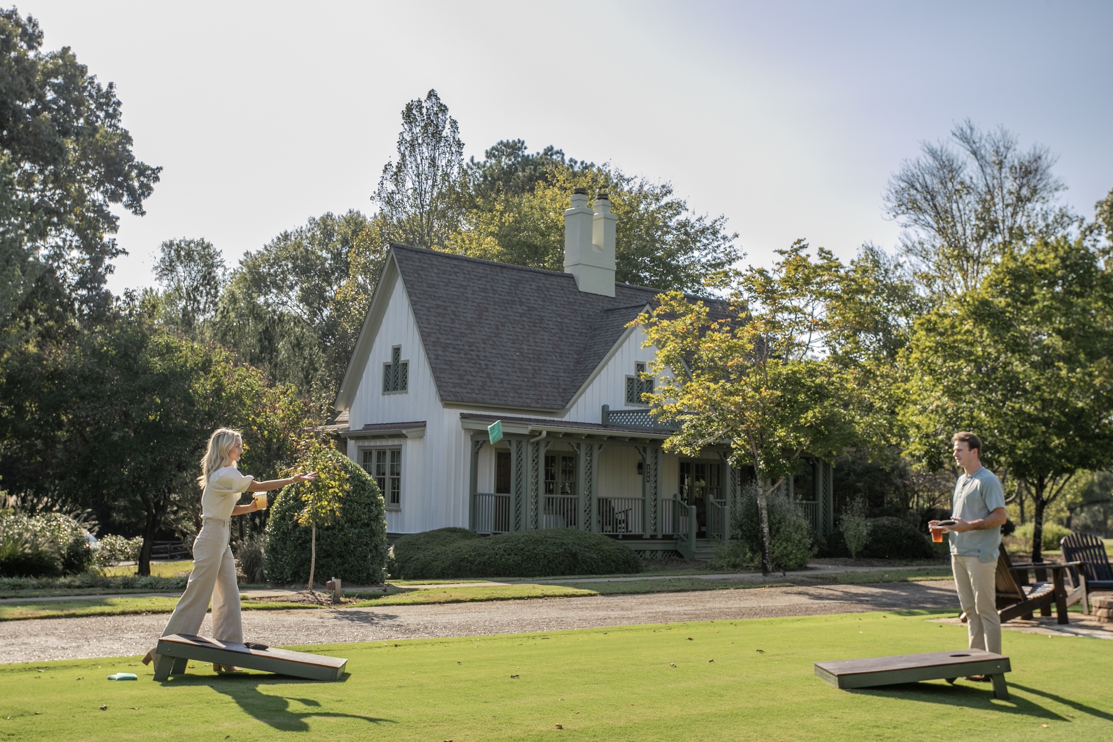 A couple playing corn hole in the Barnsley Resort village green. A cottage is in the background, it is a sunny day.