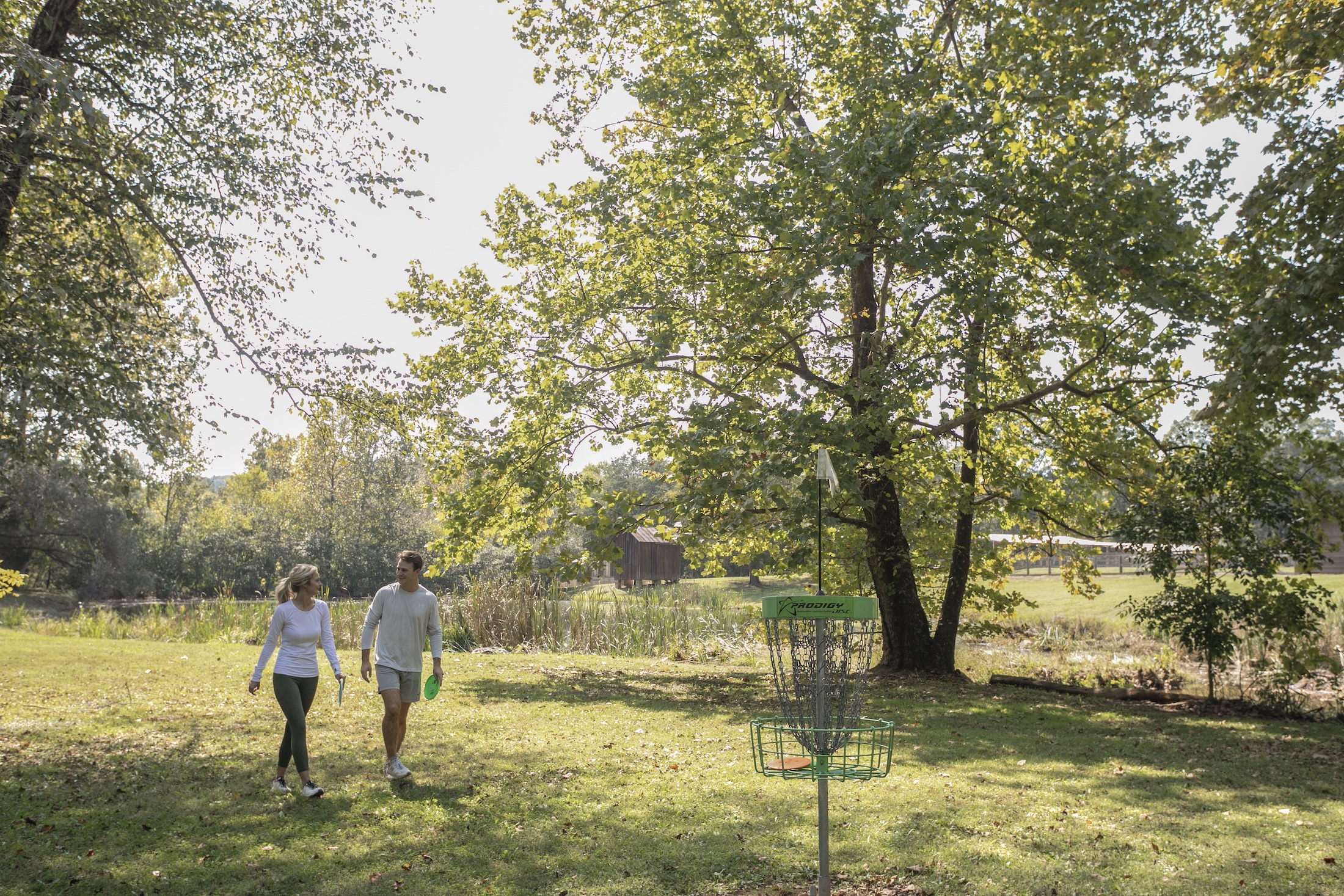 A couple walking along a disc golf course lined with trees, a green disc golf target in the foreground.