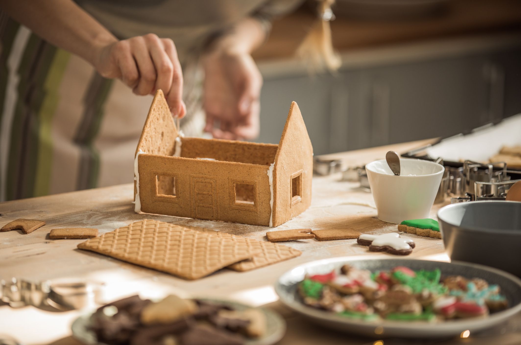 A woman making a ginger bread house on a wooden table.