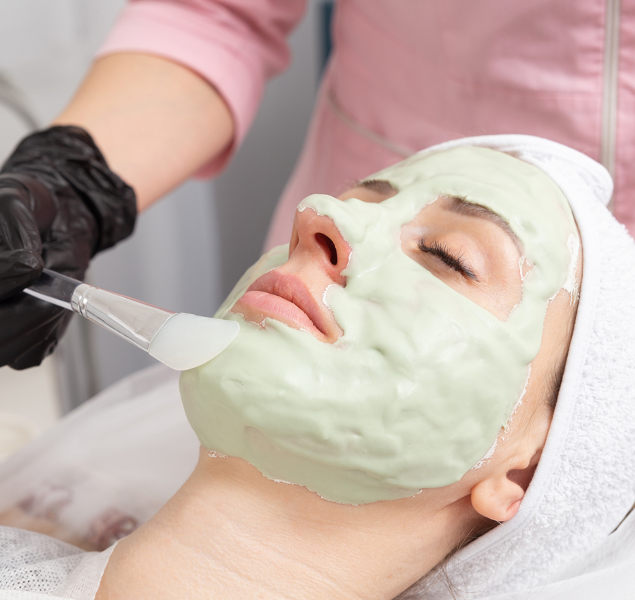 A spa worker uses a brush to apply a facial product to a woman's face. The woman is relaxed with her eyes closed.