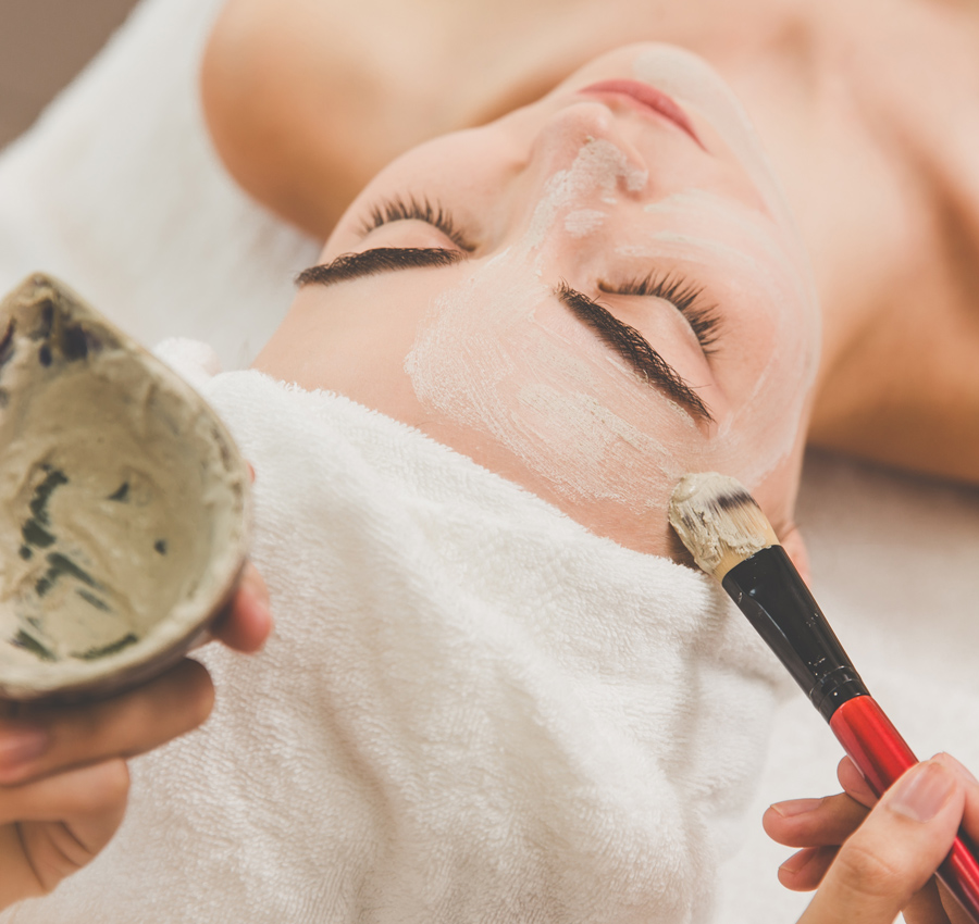 A spa worker uses a brush to apply a facial product to a woman's face. The woman is relaxed with her eyes closed.