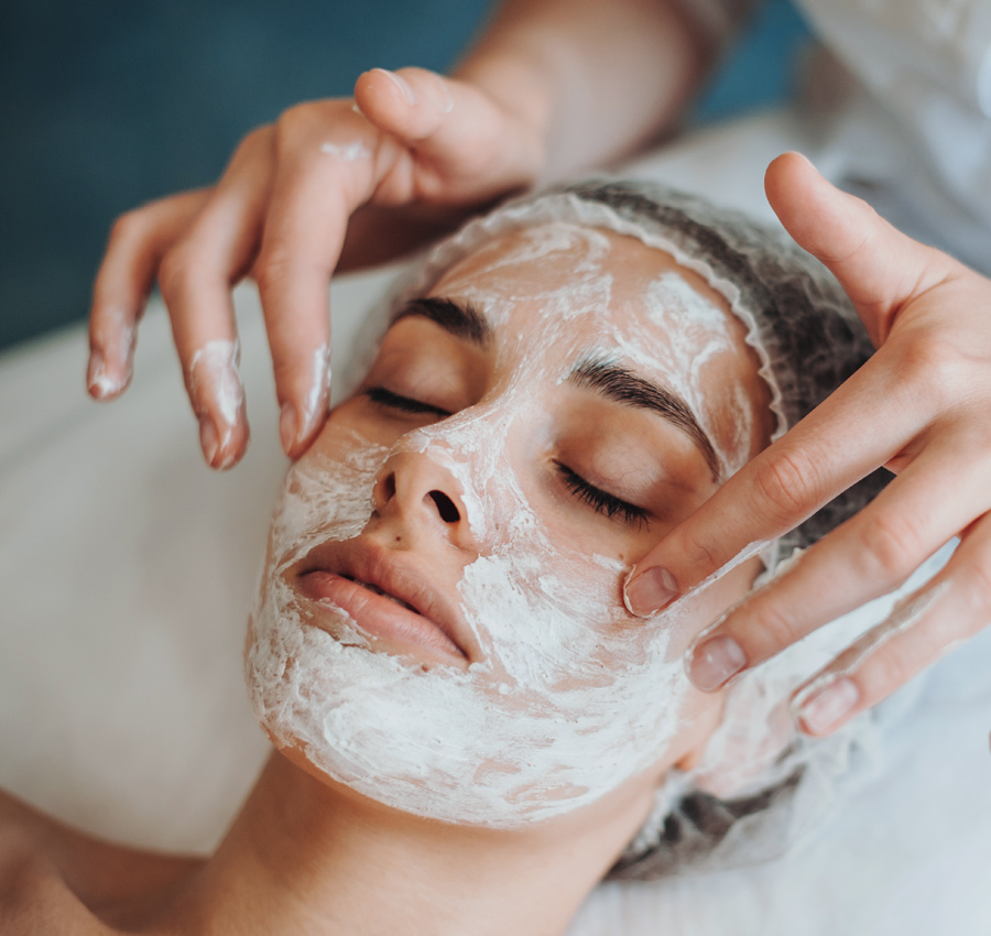 A spa worker gently applies a facial product to a woman's face with their hands. The woman is relaxed with her eyes closed.