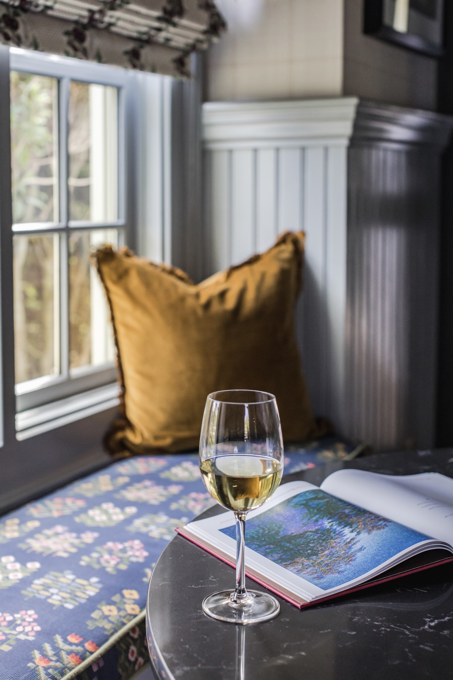 A window bench with colorful cushion and throw pillow. An open magazine and glass of white wine sit on table in front of the seat.