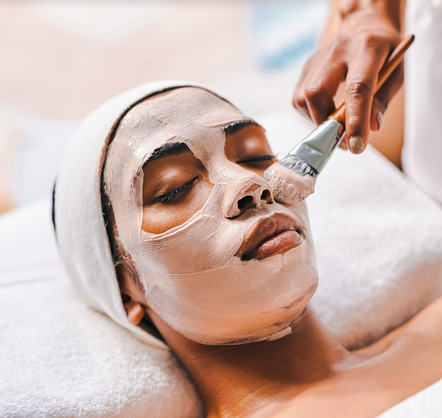 A spa worker uses a brush to apply a facial product to a woman's face. The woman is relaxed with her eyes closed.