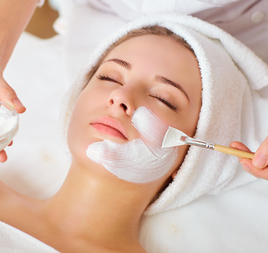 A spa worker uses a brush to apply a facial product to a woman's face. The woman is relaxed with her eyes closed.