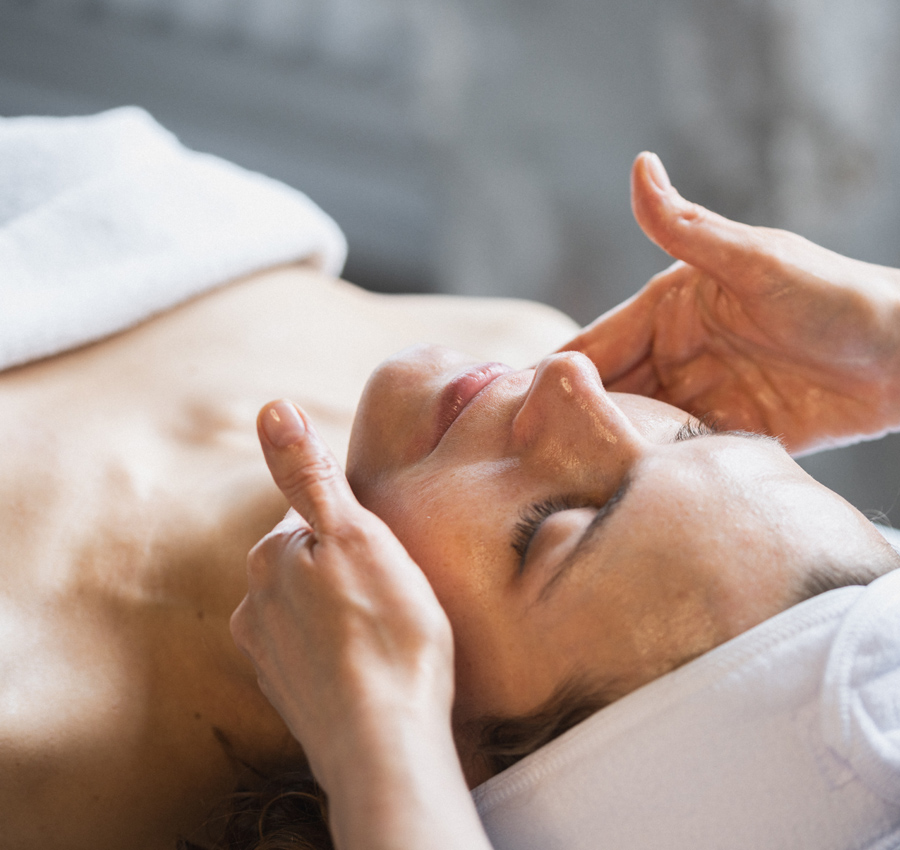 A spa worker gently applies a facial product to a woman's face with their hands. The woman is relaxed with her eyes closed.