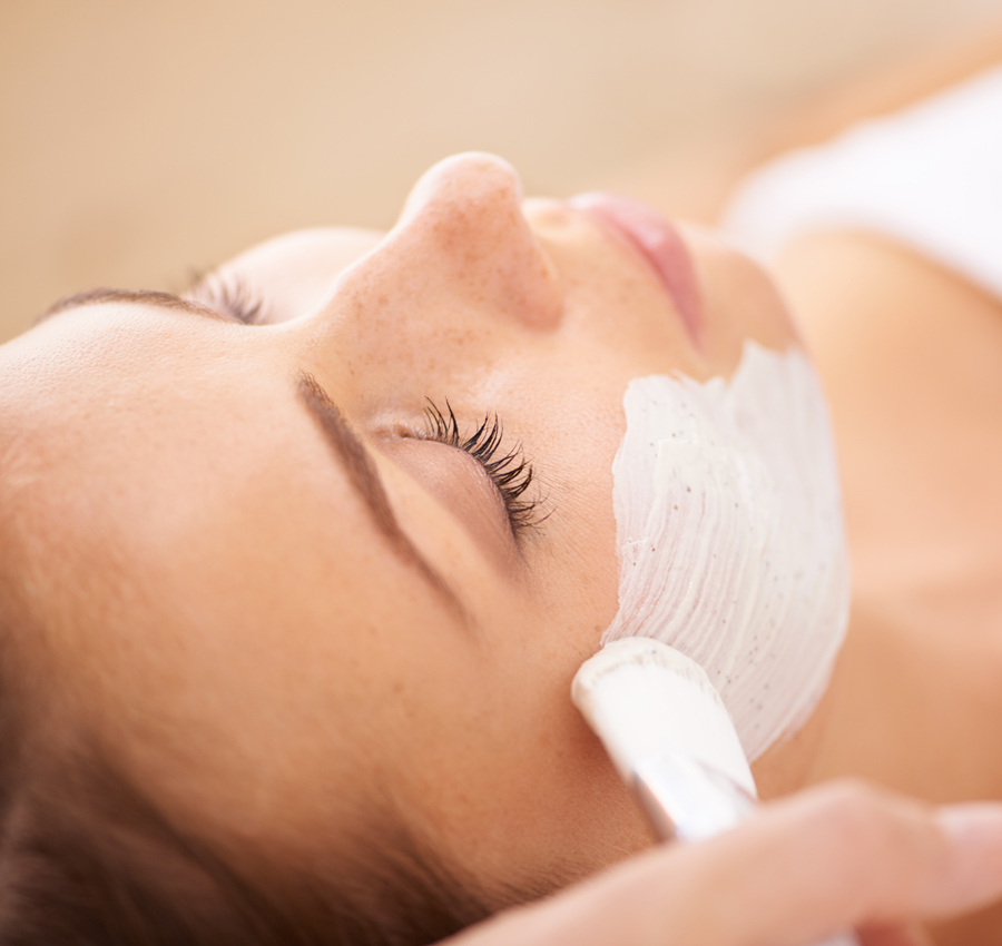 A spa worker uses a brush to apply a facial product to a woman's face. The woman is relaxed with her eyes closed.