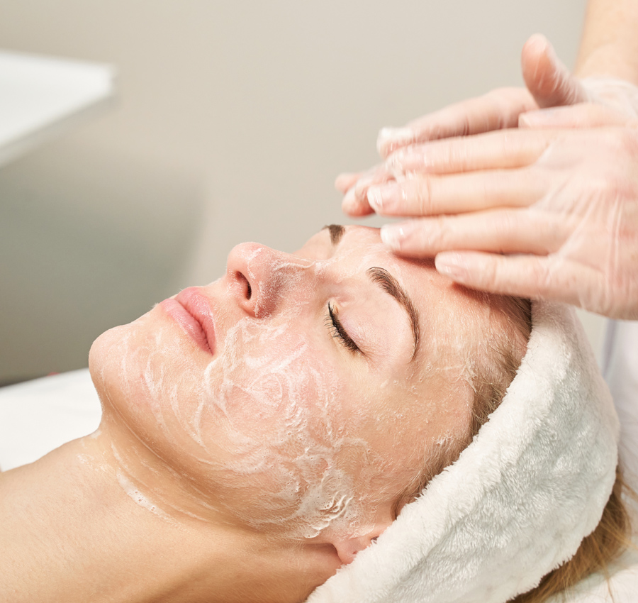 A spa worker gently applies a facial product to a woman's face with their hands. The woman is relaxed with her eyes closed.