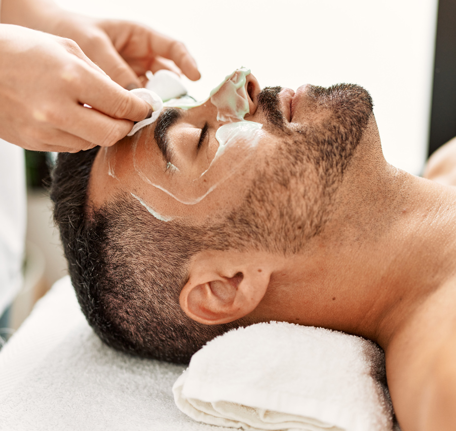 A spa worker gently applies a facial product to a man's face with their hands. The man is relaxed with his eyes closed.