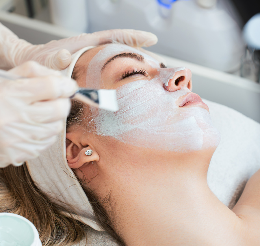 A spa worker uses a brush to apply a facial product to a woman's face. The woman is relaxed with her eyes closed.