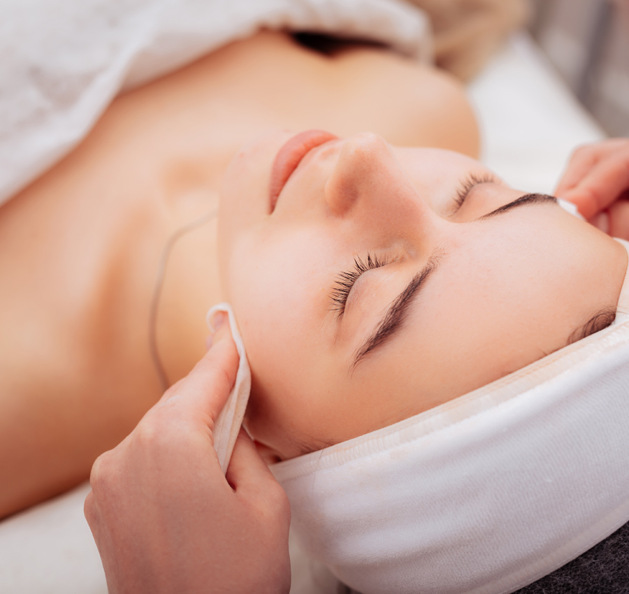 A spa worker gently applies a facial product to a woman's face with their hands. The woman is relaxed with her eyes closed.