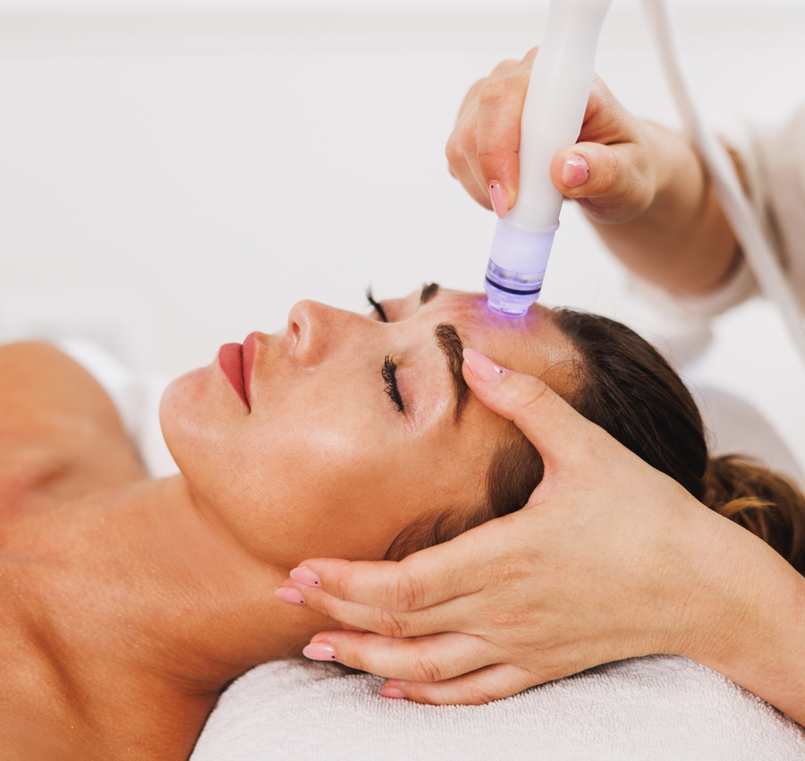 The gloved hands of a spa worker uses a HydraFacial tool on a woman's face.