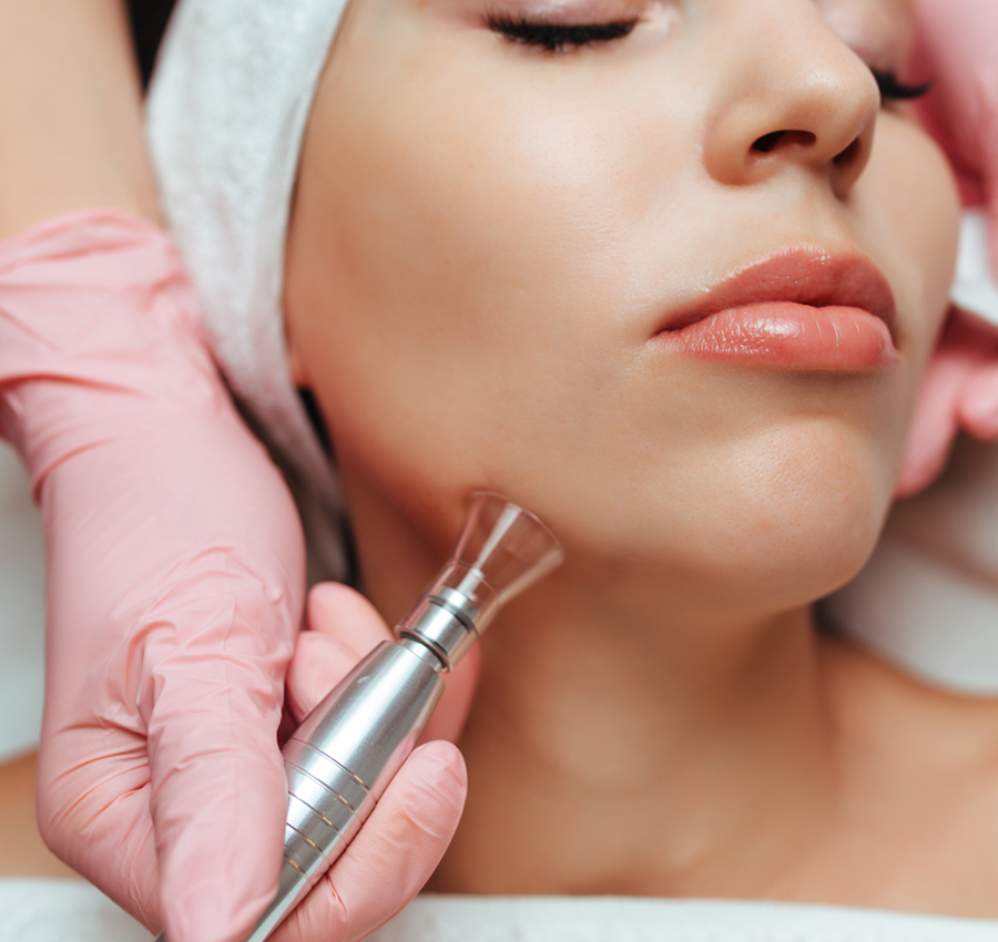 The gloved hands of a spa worker uses a HydraFacial tool on a woman's face.