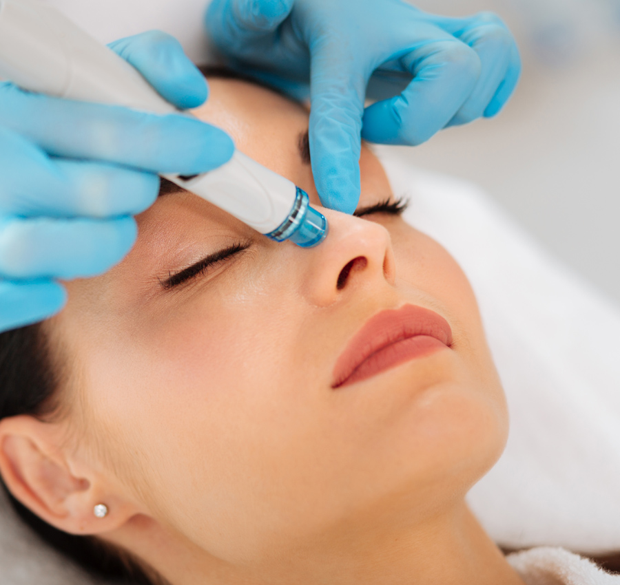 The gloved hands of a spa worker uses a HydraFacial tool on the nose of a woman.