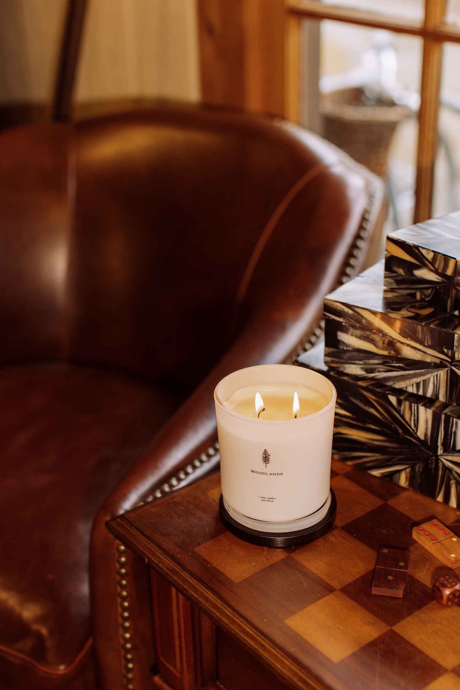 A Woodlands branded candle is lit on a wooden table next to a leather chair.