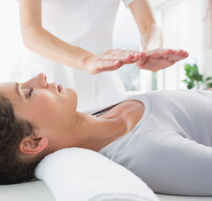 A woman is lying on a spa bed and receives an energy treatment from the spa worker who is holding their hands above the woman's chest.