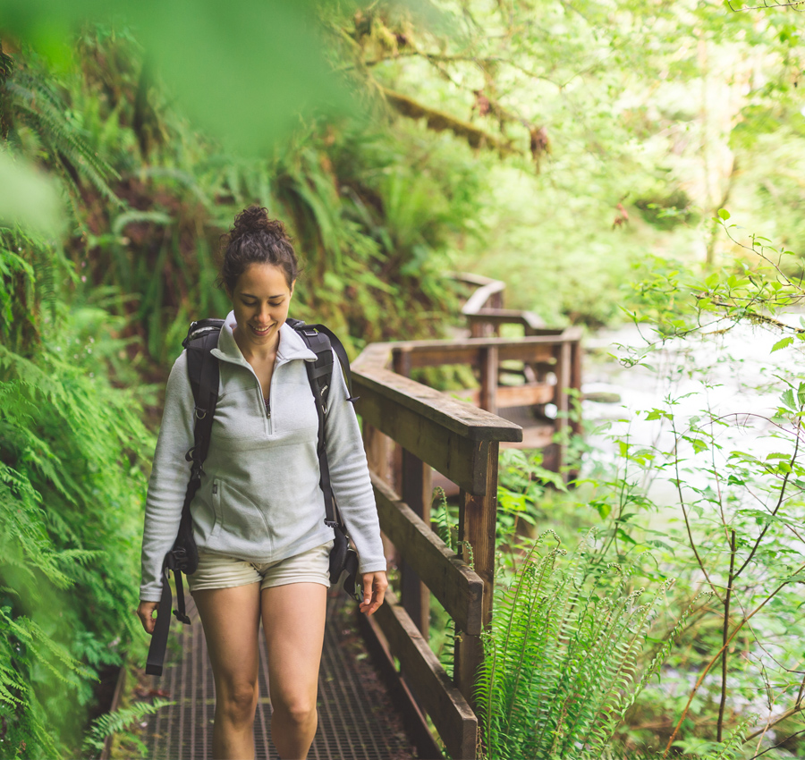 A woman walks along a wooden bridge next to a river in the woods.