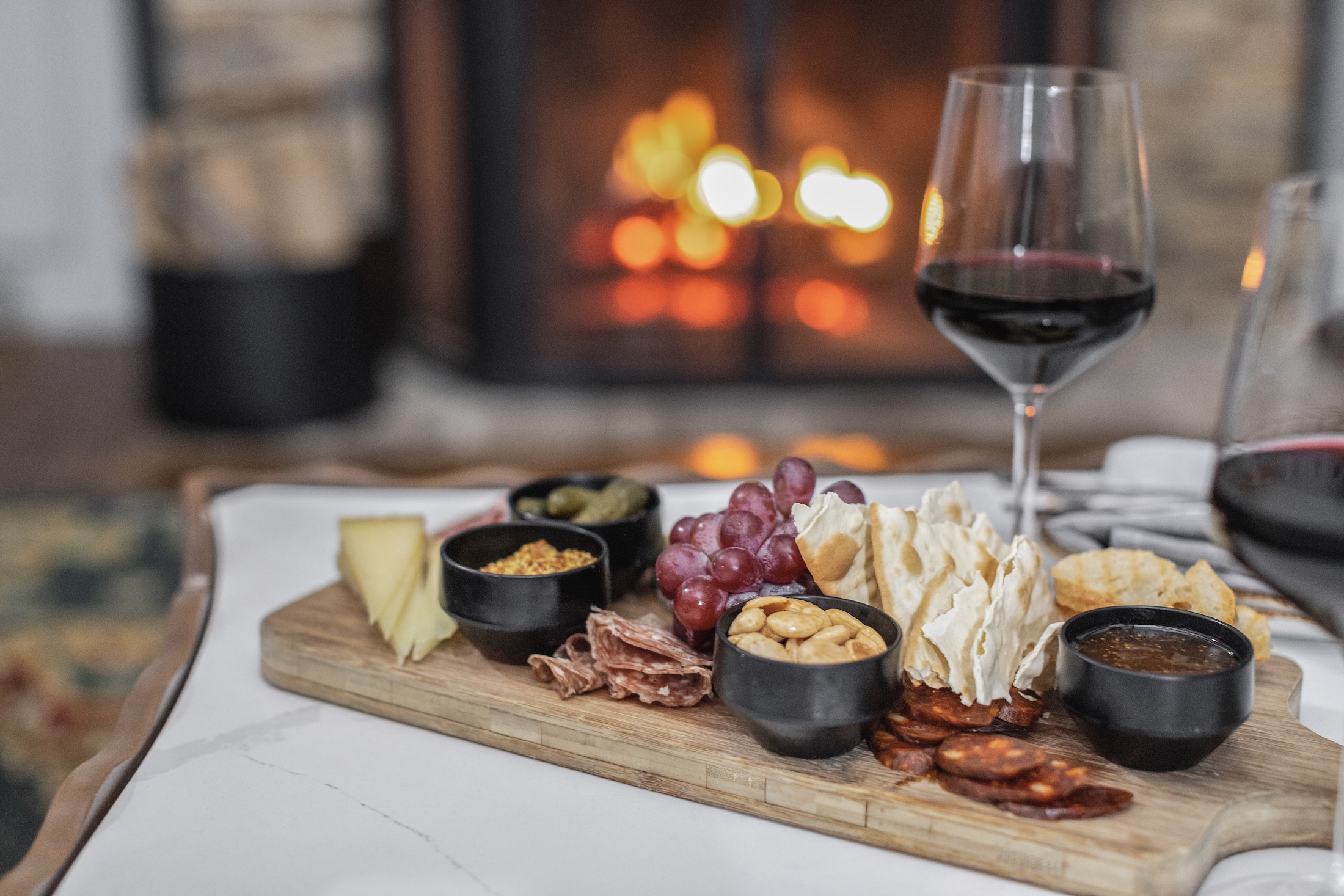 a charcuterie board filled with grapes, pickles, variety of cheeses, meats and crackers with a side of a glass of red wine