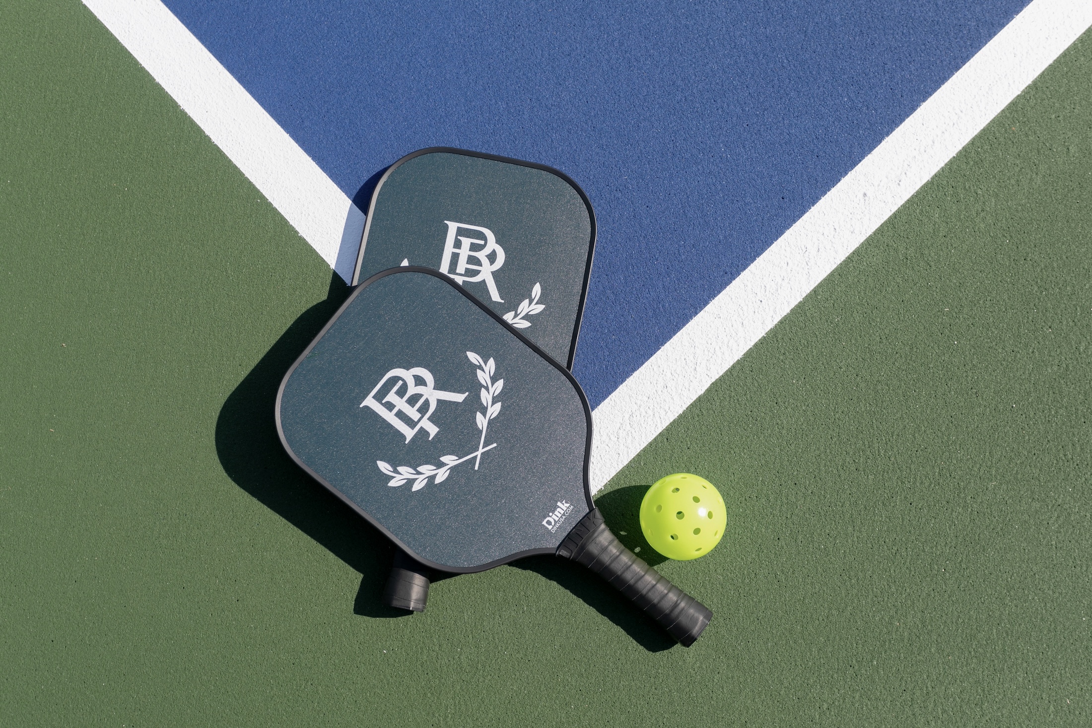 Two pickleball paddles with the Barnsley Resort monogram are laid one on-top the other with a pickleball on the corner of a pickleball court.