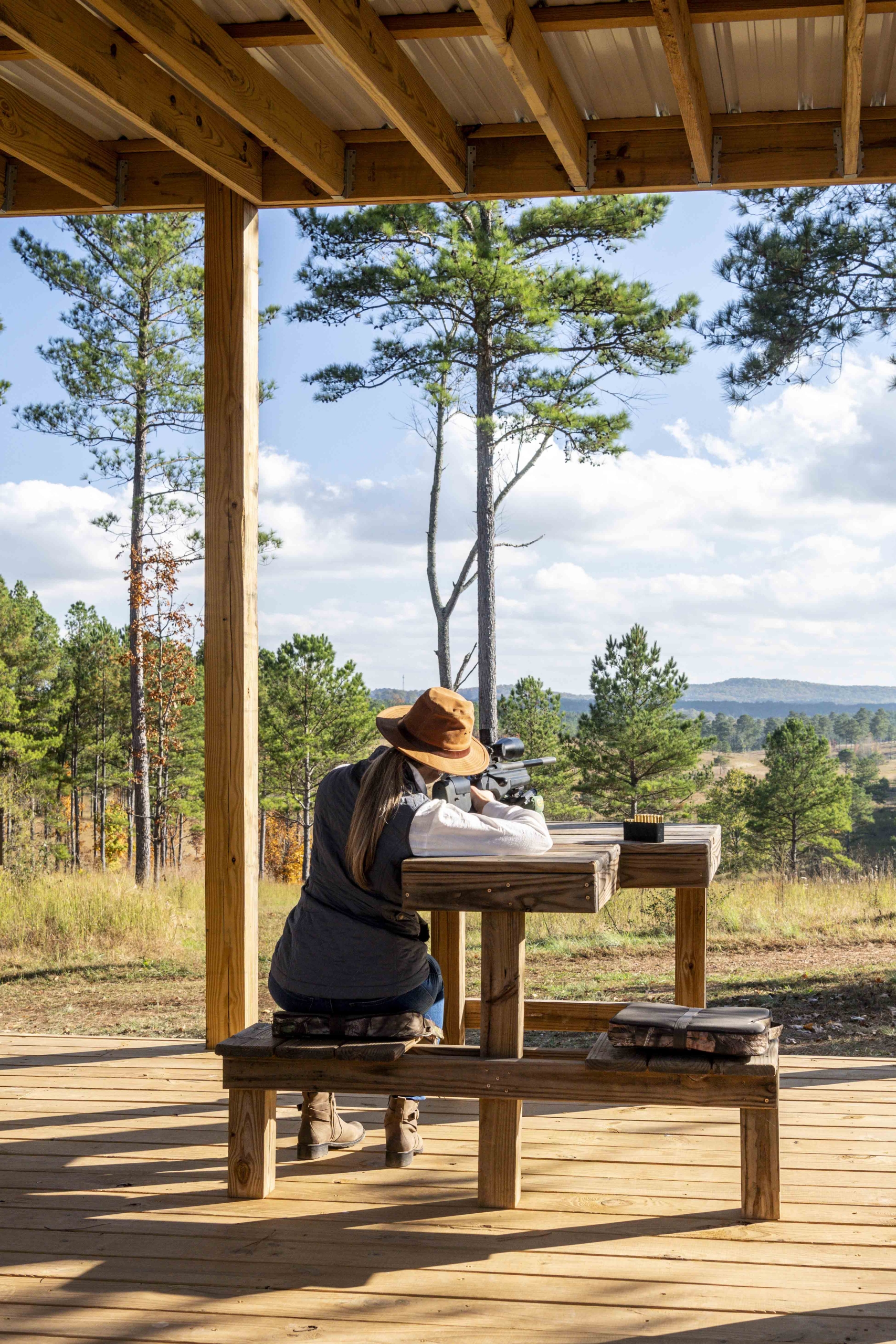 A woman wearing a wide brimmed hat sits on a covered, wooden platform as she aims a hunting rifle at a target in the distance.