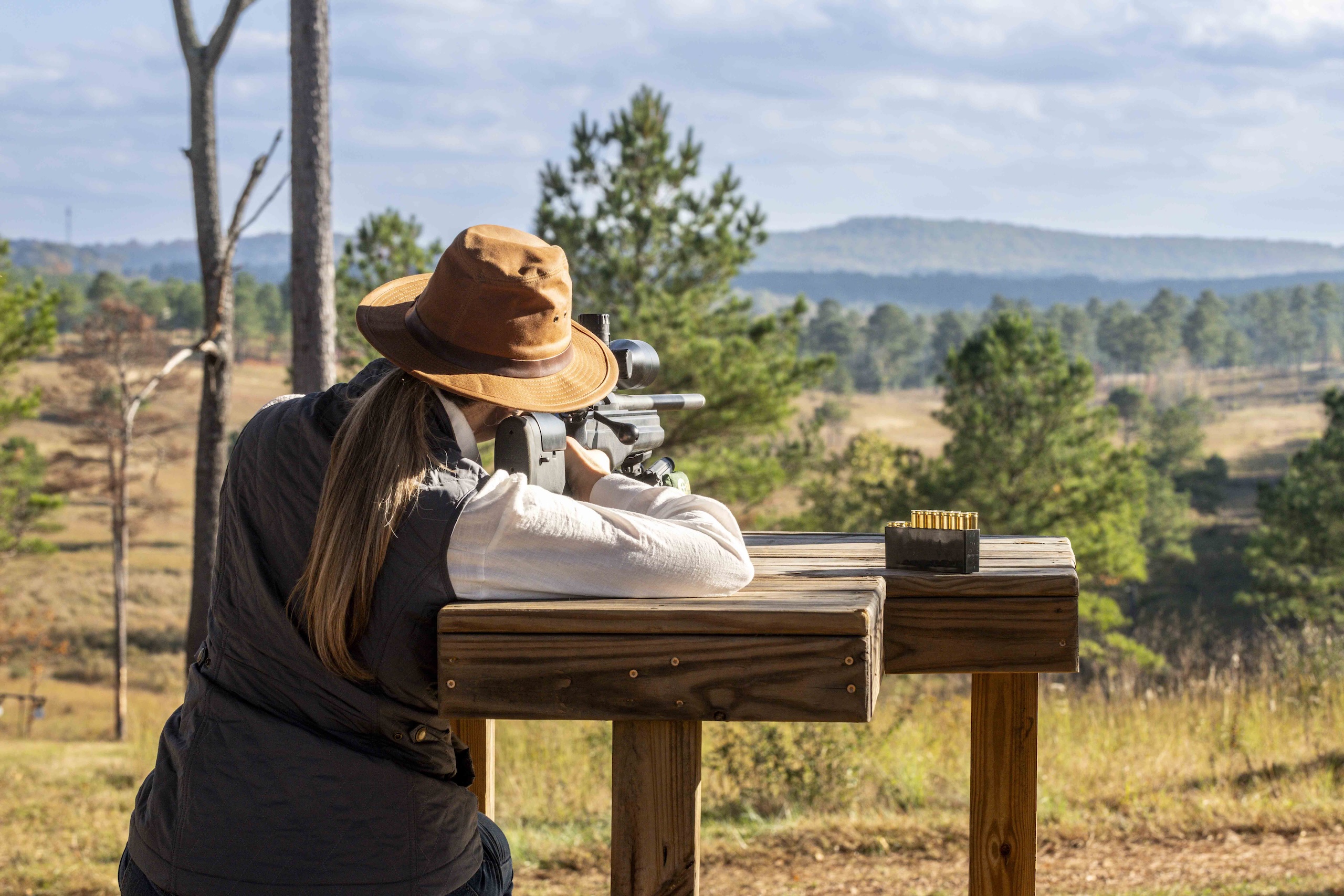 A woman wearing a wide brimmed hat leans up against a wooden platform as she aims a hunting rifle at a target in the distance.