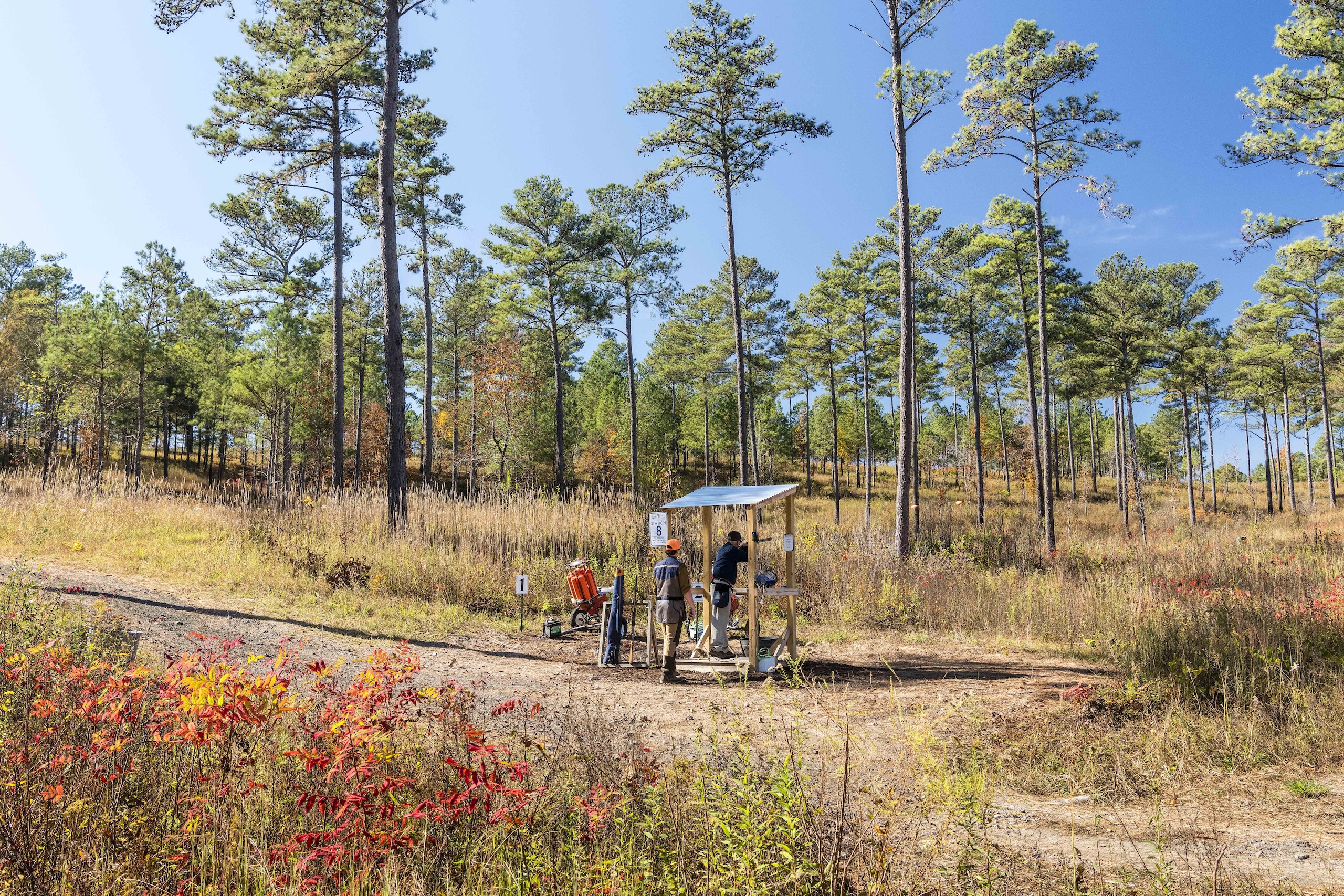 A man shooting a rifle at a sporting clay station while a worker watches.