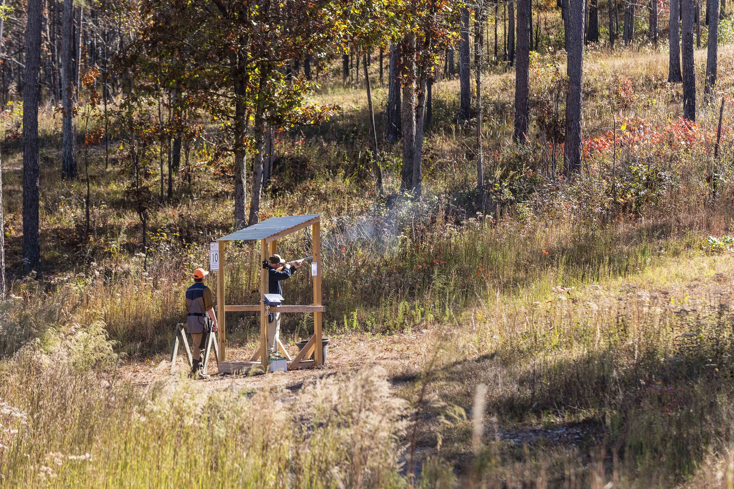 A man shooting a rifle at a sporting clay station while a worker watches.