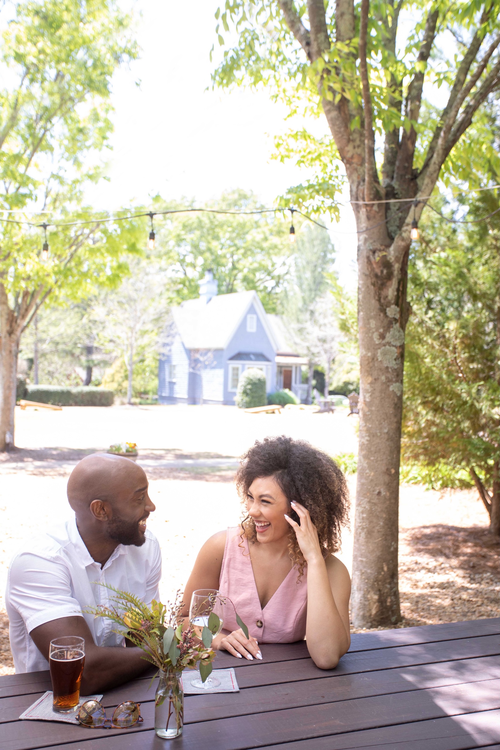 A couple enjoying a beer and a wine sitting on a picnic bench under trees on a sunny day at Biergarten.