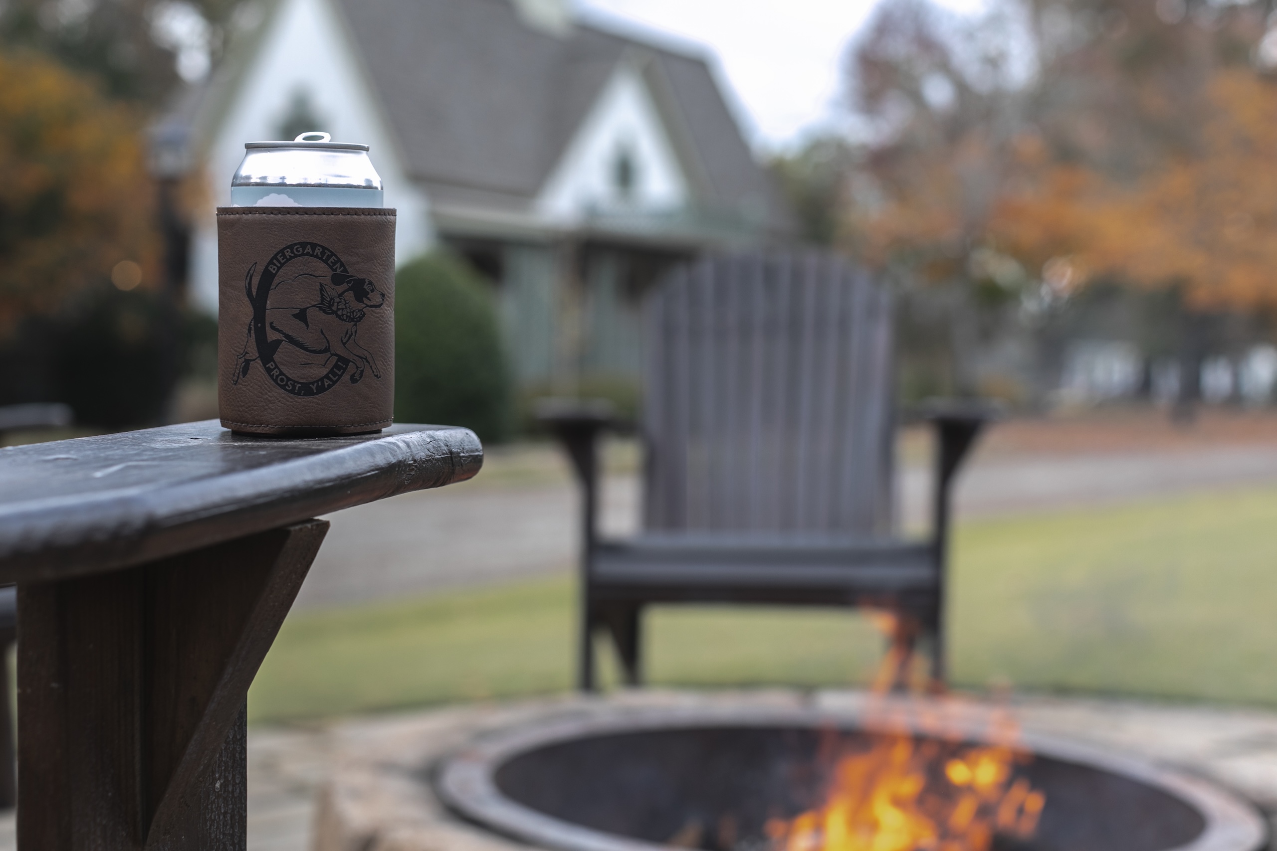 A can of beer with a Biergarten branded koozie sits on the arm of an adirondack chair in front of a fire pit with a fire burning in it. Orange leaved trees and a cottage can be seen in the background.