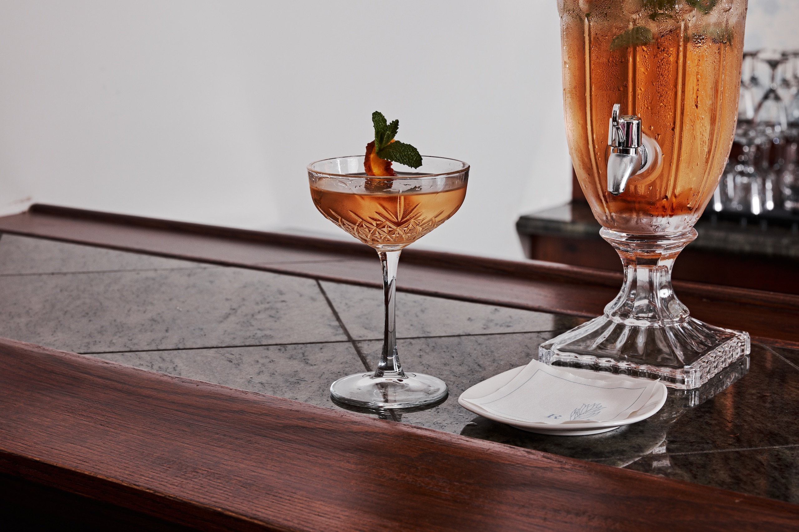 An amber colored cocktail in a crystal coupe glass sitting on a marble and dark wooden counter top.