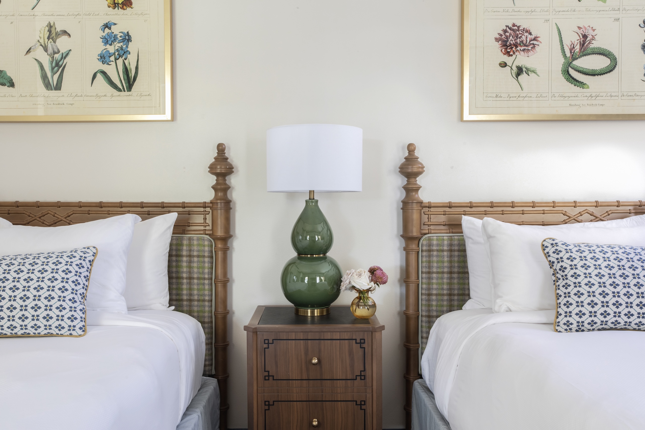 Two queen beds on either side of a night table with a green lamp and small bouquet of flowers.